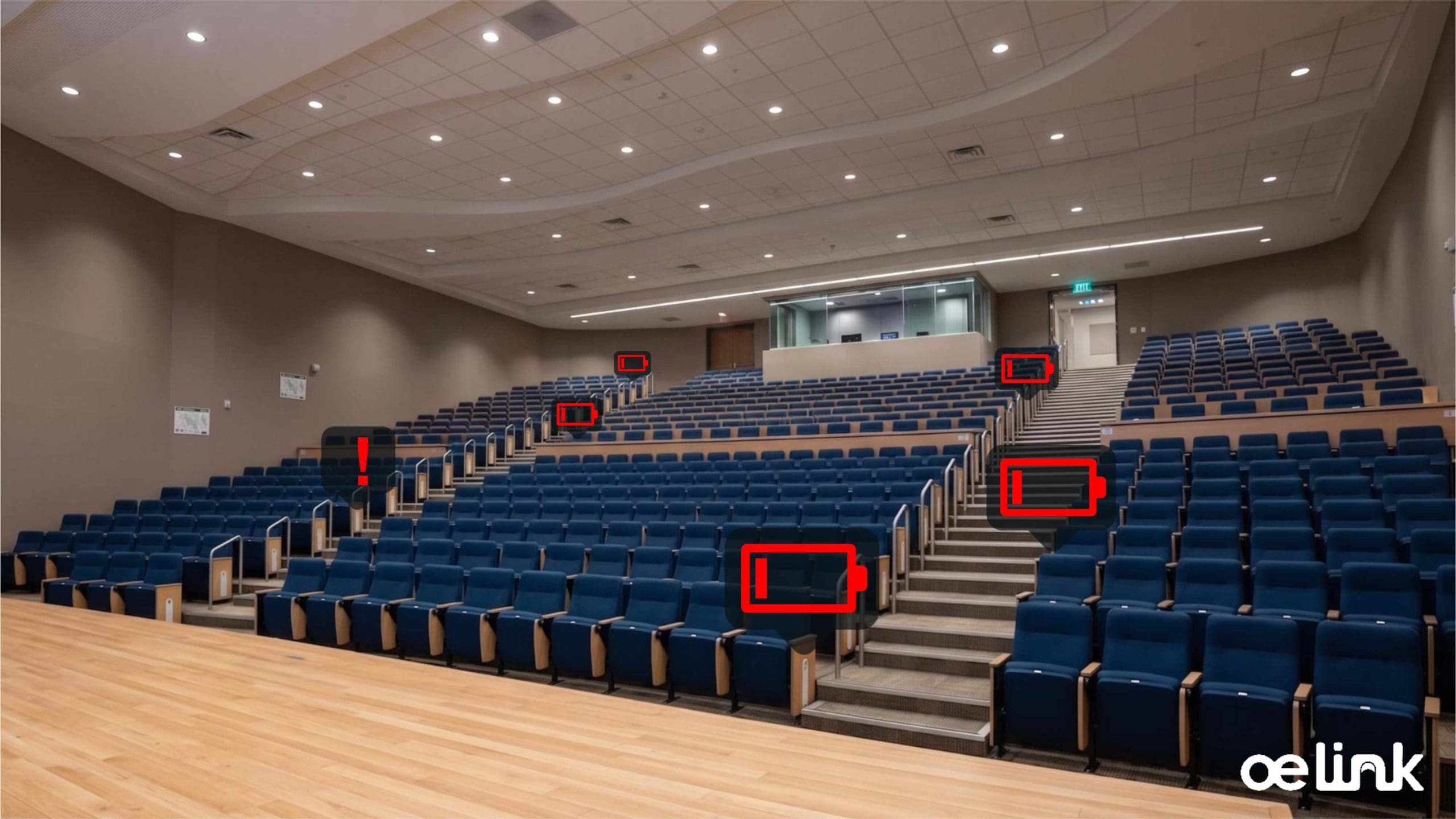 Empty auditorium with curved rows of blue seats, a wooden stage foreground, and a glass-backed control booth at the rear.