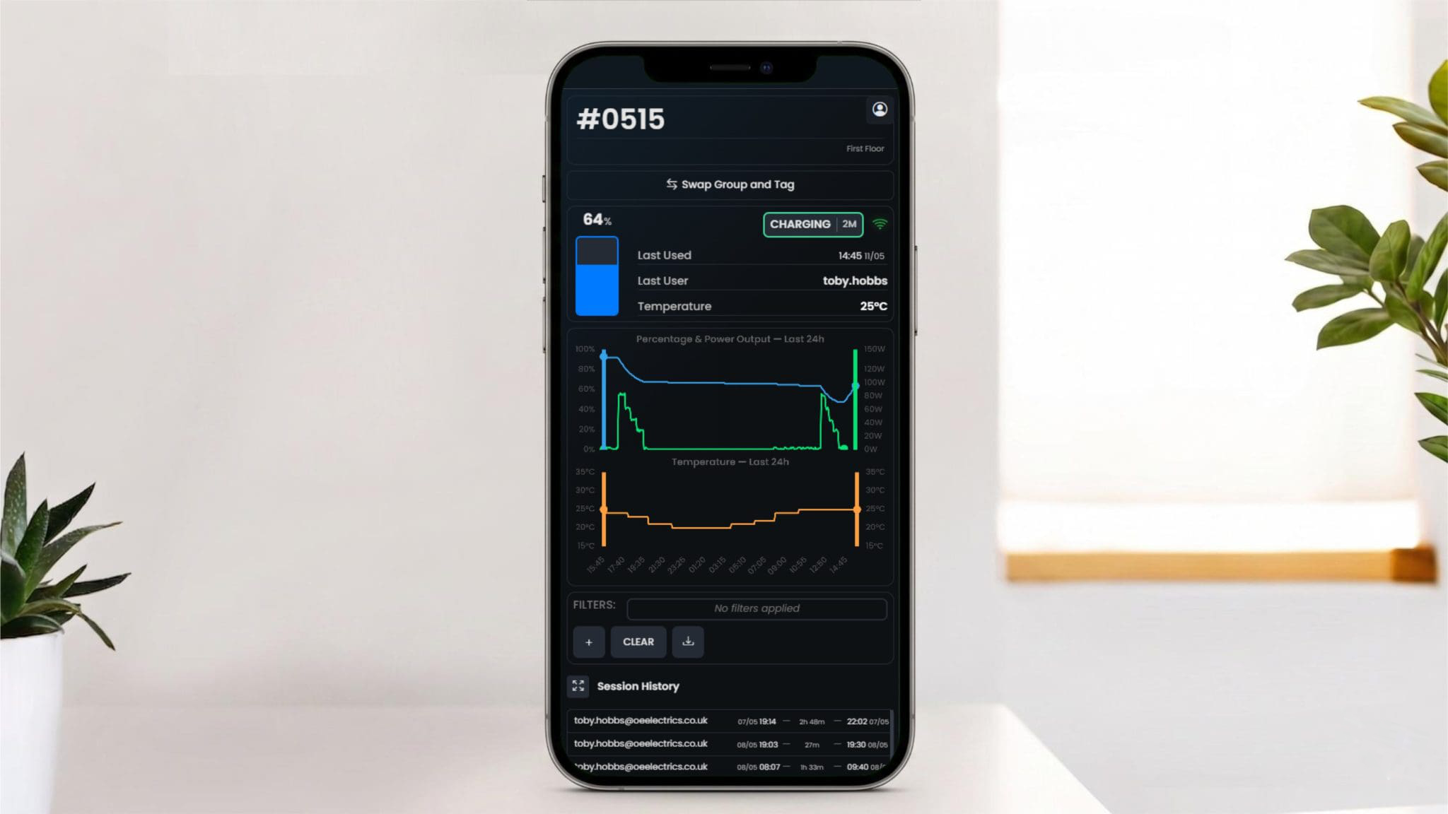 Close-up of a smartphone displaying a dark monitoring dashboard with battery, temperature, and usage graphs on a desk with plants nearby.