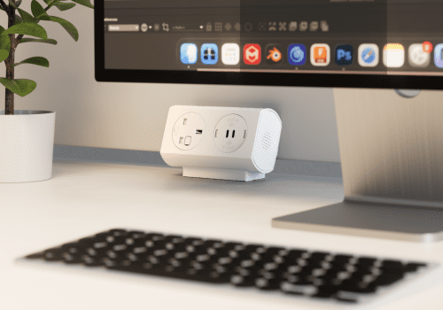 White power strip on a desk next to a computer monitor, with a potted plant to the left.