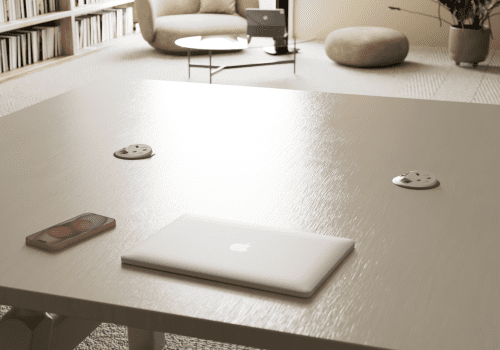Wide view of a cozy home workspace: a closed MacBook and an iPhone on a wooden table, with two recessed outlet plates, a bookshelf, and a seating area in the background.