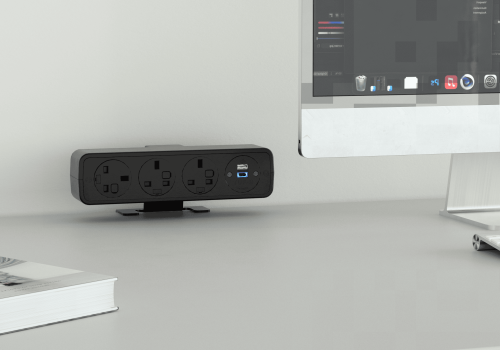 Black power strip with three outlets and a USB port on a desk beside a computer monitor and keyboard