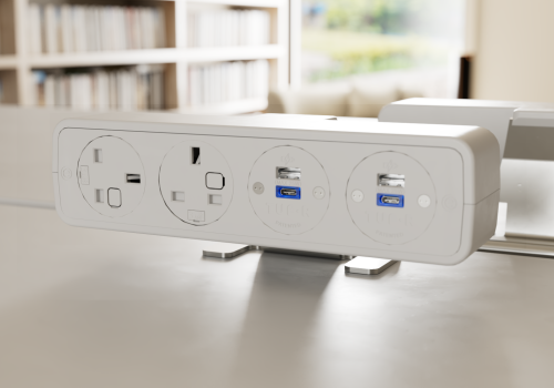 White power hub on a desk featuring three outlets and two USB ports (blue USB ports) for devices, with a blurred bookshelf and window in the background.